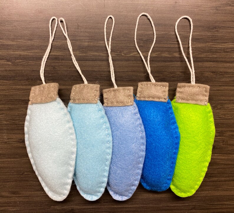 May include: Five handmade felt Christmas light ornaments in shades of blue and green, with tan tops and white string loops for hanging. The ornaments are arranged in a row against a dark wood grain background.