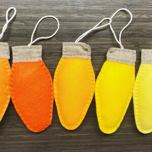 May include: Five felt ornaments shaped like light bulbs in shades of orange and yellow. The ornaments have a white string attached to the top for hanging.