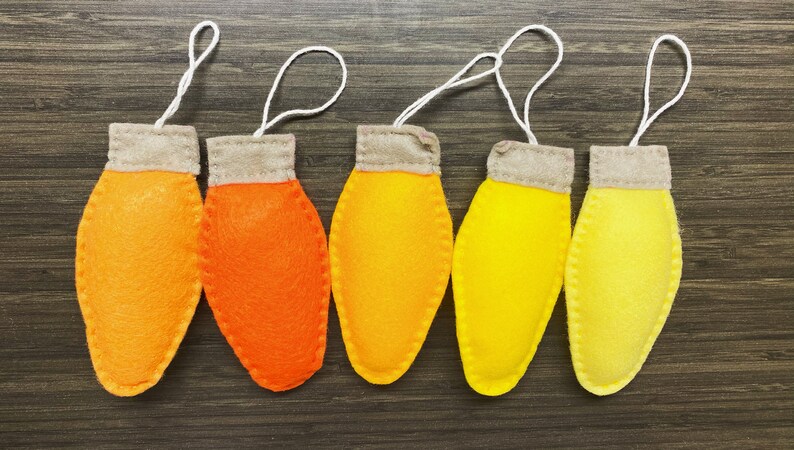 May include: Five felt ornaments shaped like light bulbs in shades of orange and yellow. The ornaments have a white string attached to the top for hanging.