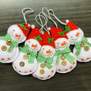 May include: A collection of six snowman ornaments. Each snowman is white with a red hat, orange carrot nose, and a green and white checkered scarf. They have wooden button accents and red star details.