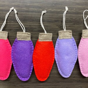 May include: Five felt Christmas light ornaments in shades of pink, purple, and red, each with a gray top and a white string for hanging. The ornaments are arranged in a row on a dark wood surface.