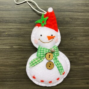 May include: A white felt snowman Christmas ornament with a red hat, green star, and a green and white checkered bow. The snowman has a carrot nose, a smiling mouth, and two wooden buttons. A small silver bell is attached to the bow.