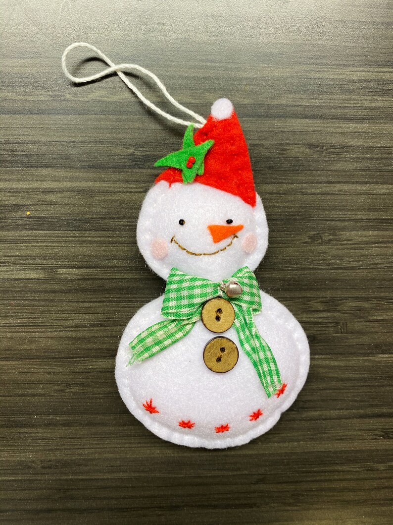 May include: A white felt snowman Christmas ornament with a red hat, green star, and a green and white checkered bow. The snowman has a carrot nose, a smiling mouth, and two wooden buttons. A small silver bell is attached to the bow.
