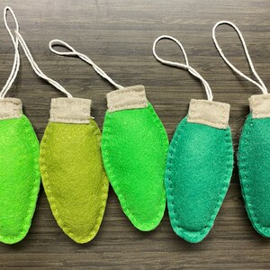 May include: Five handmade felt Christmas light ornaments in shades of green. Each ornament has a tan top and a white string for hanging. The ornaments are arranged on a dark wood grain surface.