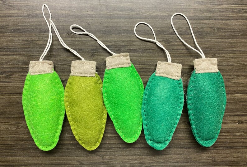 May include: Five handmade felt Christmas light ornaments in shades of green. Each ornament has a tan top and a white string for hanging. The ornaments are arranged on a dark wood grain surface.