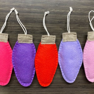 May include: Five felt ornaments shaped like light bulbs, hanging from white strings. The ornaments are pink, purple, red, lavender, and light pink.