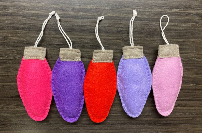 May include: Five felt ornaments shaped like light bulbs, hanging from white strings. The ornaments are pink, purple, red, lavender, and light pink.