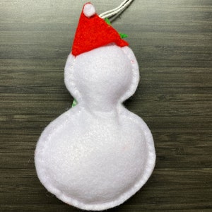 May include: A handmade snowman ornament made of white felt, wearing a red Santa hat with a white pom-pom. The ornament has a white string for hanging and is designed for Christmas decorations.