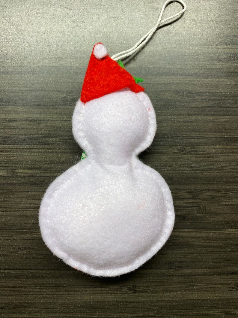 May include: A handmade snowman ornament made of white felt, wearing a red Santa hat with a white pom-pom. The ornament has a white string for hanging and is designed for Christmas decorations.