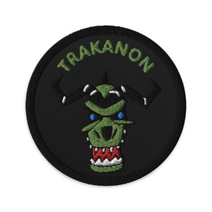 May include: A black and green embroidered patch with a cartoon monster face and the text "TRAKANON" around the edge.