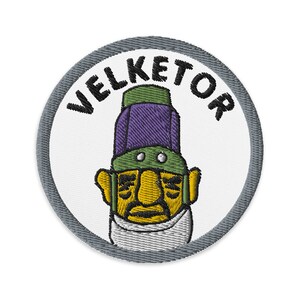 May include: A round, gray embroidered patch with the word "VELKETOR" in black lettering around the edge. In the center is a cartoon character with a purple and green hat, a yellow face, and a white beard.