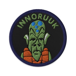 May include: A black and blue embroidered patch with a green alien wearing an orange life vest. The text "INMORUUK" is written in a circle around the alien.