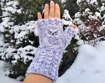 May include: A light gray knitted fingerless glove with an owl design. The owl has yellow eyes and is centered on the glove. The glove is made of a textured yarn and is set against a snowy background.