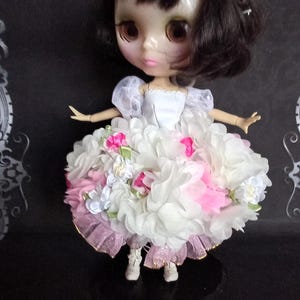 Può includere: Una bambola con capelli castano scuro e una fascia bianca, che indossa un corpetto bianco con maniche a sbuffo trasparenti e una gonna voluminosa coperta da strati di fiori in tessuto bianco e rosa, con orlo a balze rosa e stivaletti bianchi.