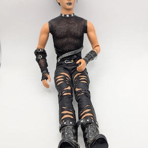 May include: A posable doll with short dark hair and dark makeup, wearing a black mesh sleeveless top, a studded belt, and black distressed pants with multiple horizontal rips. The doll features black arm cuffs, a studded choker, and black boots.