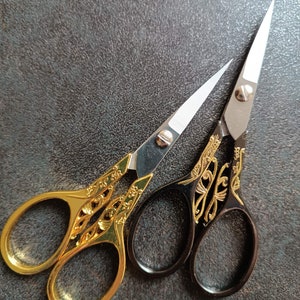 Puede incluir: Dos pares de tijeras decorativas con mangos dorados y negros. Un par tiene mangos dorados con un diseño floral y el otro par tiene mangos negros con un diseño floral dorado.
