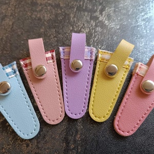 May include: Six colorful leather cases with a checkered fabric lining and a silver button closure. The cases are in shades of blue, pink, purple, yellow, and light pink.