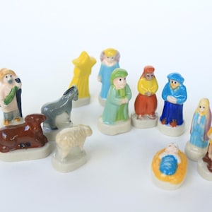 May include: A collection of small, colorful ceramic figurines depicting a nativity scene. The set includes figures of Mary, Joseph, baby Jesus, shepherds, animals, and the three wise men. The figurines are painted with bright colors and set on small, white bases.
