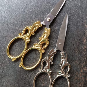 May include: Two pairs of decorative scissors with ornate gold and silver handles. The silver blades contrast with the detailed, swirling handle design. These scissors are likely for crafting or decorative use.