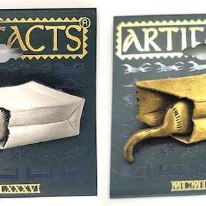 May include: Two metal pins, one silver and one gold, each depicting a cat's tail protruding from a paper bag. The pins are on dark blue cards with the word "ARTIFACTS" in gold. The Roman numeral "MCMLXXXVI" is at the bottom.