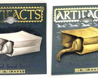 May include: Two metal pins, one silver and one gold, each depicting a cat's tail protruding from a paper bag. The pins are on dark blue cards with the word "ARTIFACTS" in gold. The Roman numeral "MCMLXXXVI" is at the bottom.