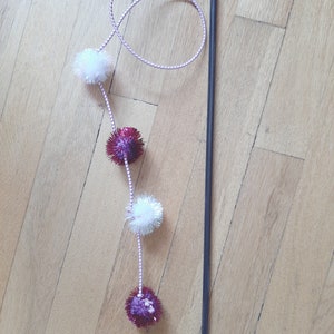 May include: A black wand with a pink and white string attached to it. The string has four pom-poms, two white and two red with silver glitter.