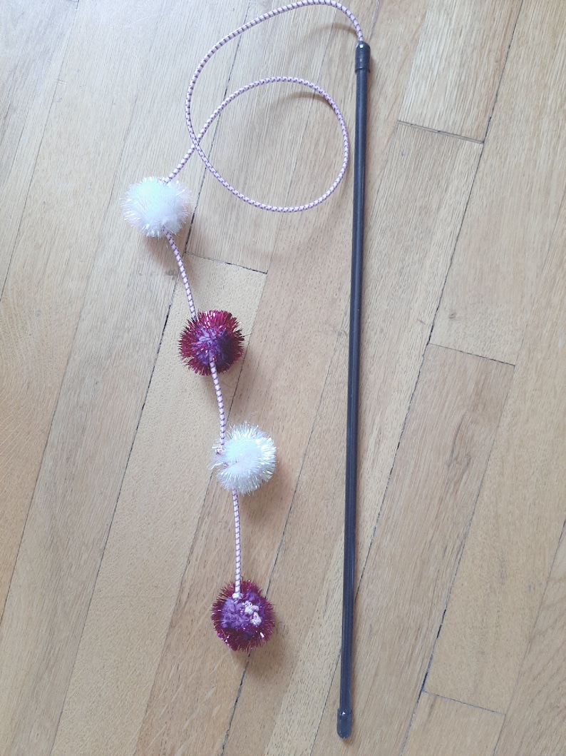 May include: A black wand with a pink and white string attached to it. The string has four pom-poms, two white and two red with silver glitter.