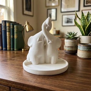 Puede incluir: Una figura blanca de elefantes con las trompas entrelazadas, sobre una base redonda. La escultura es un elemento decorativo, probablemente para un hogar u oficina. Los elefantes se enfrentan, formando un corazón con sus trompas. La base mide unos 15 cm de diámetro.