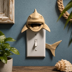 May include: A light beige shark-shaped light switch cover plate featuring a sculpted shark head with an open mouth at the top and a shark tail fin extending from the right side, mounted over a standard white rectangular toggle light switch.