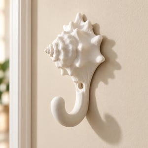 Puede incluir: Un gancho de pared blanco mate con forma de caracola. El diseño presenta la textura intrincada y en espiral de una concha marina con un gancho curvo que se extiende desde la parte inferior. Un orificio de montaje circular es visible en la base.