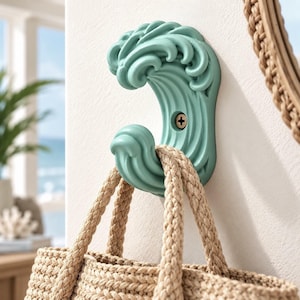May include: A wall-mounted hook shaped like a stylized ocean wave in a matte seafoam green finish. The hook features a curved design with deep ridges and is secured to the wall with a single visible screw. A beige woven rope bag hangs from the hook.