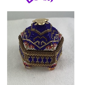 Può includere: Una piccola scatola esagonale di perline con un design dorato e viola. Il coperchio della scatola è a forma di rospo ed è anche perlato. La scatola è appoggiata su una superficie bianca.