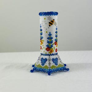 May include: A white beaded vase with a blue rim and base. The vase is decorated with a bee, flowers, and leaves in various colors.