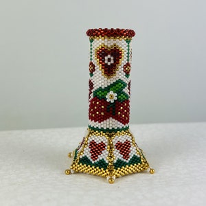 Puede incluir: Un jarrón de cuentas con un patrón de fresas y flores rojas, blancas y verdes. El jarrón tiene una base hexagonal con cuentas doradas.