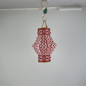 May include: A red and white beaded lantern ornament with a gold trim. The lantern is hanging from a gold hook with a green bead.