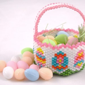 May include: A small pink and white beaded Easter basket filled with pastel coloured candy eggs. The basket has a beaded handle and a green base. The candy eggs are scattered around the basket.