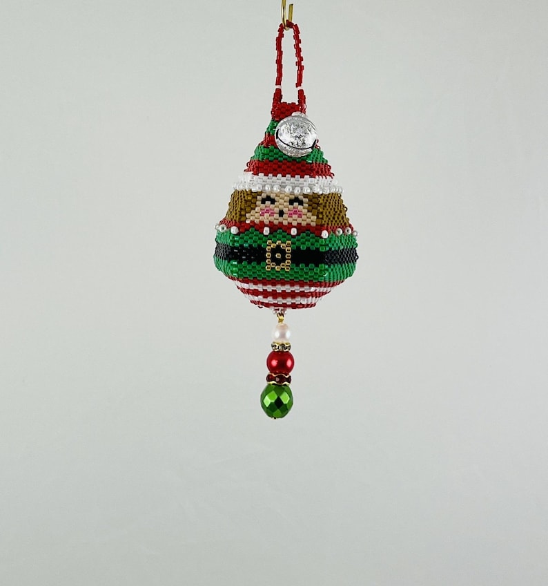 May include: A beaded Christmas ornament shaped like an elf, with a red and white striped hat, green and black striped body, and a gold belt. The elf has a white face with brown hair and a silver bell hanging from the hat.