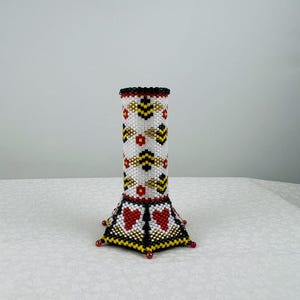 May include: A white, black, gold and red beaded vase with a geometric pattern. The vase has a cylindrical body and a hexagonal base.
