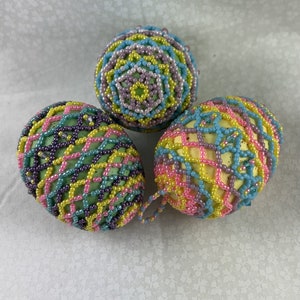May include: Three decorative Easter eggs. Each egg is covered in colorful beads. The eggs are in shades of yellow, pink, blue, green, and purple. The eggs are on a white background.