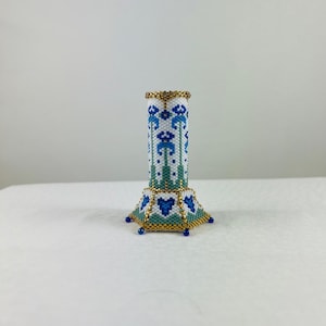 May include: A small, white, blue, and gold beaded vase with a geometric design. The vase has a cylindrical body and a hexagonal base.