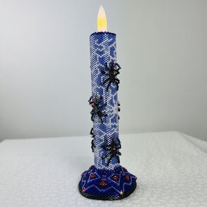May include: A blue beaded candle with a white geometric pattern and black spider embellishments. The candle has a decorative base with red accents and a flickering LED flame. A unique Halloween or holiday decoration.