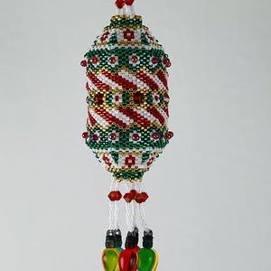 Könnte beinhalten: Ein perlenbesetzter Weihnachtsschmuck mit zylindrischer Form. Der Schmuck weist ein Spiralmuster aus roten und weißen Perlen auf, akzentuiert mit Grün und Gold. Drei herabhängende Stränge enden mit bunten Glühbirnen-Anhängern.