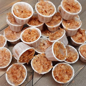 May include: A collection of small, white, fluted paper cups filled with a textured, light brown, flaky substance. The cups are arranged in a cluster, each containing a portion of the golden-brown, thin, irregular pieces that appear to be layered.