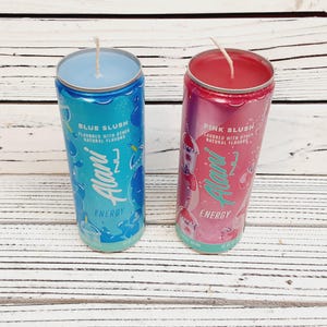 May include: Two beverage can-shaped candles. One is blue with "Blue Slush" text, the other is pink with "Pink Slush" text. Both candles have the "Alani Energy" logo and a wick.