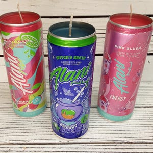 May include: Three Alani Energy drink can candles. The cans are Cherry Twist, Witch's Brew, and Pink Slush. Each can has a wick and is filled with wax. The cans are on a white wooden surface.