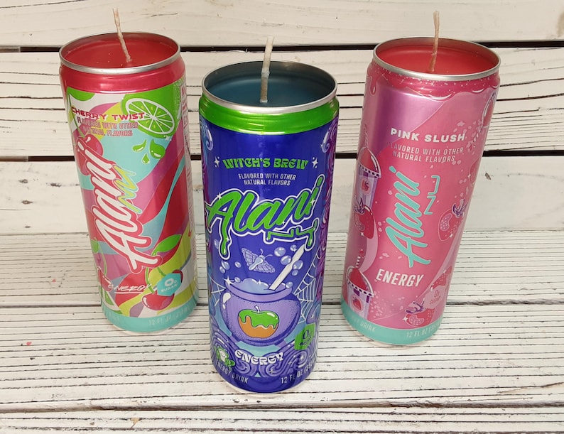 May include: Three Alani Energy drink can candles. The cans are Cherry Twist, Witch's Brew, and Pink Slush. Each can has a wick and is filled with wax. The cans are on a white wooden surface.