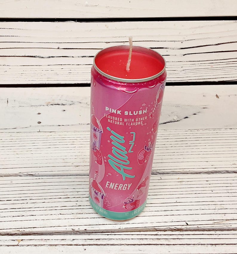 May include: A pink candle in a repurposed Alani Nu Energy drink can. The can features the words "Pink Slush" and "Energy" in a stylized font. The candle wax is a vibrant pink, and a white wick is centered.