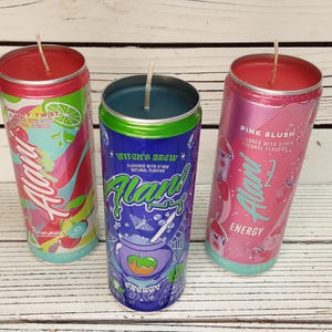 May include: Three beverage can-shaped candles. The candles are in pink, blue, and red cans with the brand name "Alani" and various flavor names. The "Cherry Twist" candle has a red top, the "Witch's Brew" candle has a blue top, and the "Pink Slush" candle has a pink top.