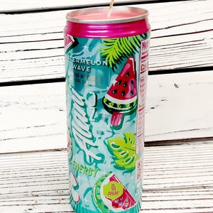 May include: A watermelon-scented candle in a repurposed 12 fl oz (355ml) can. The can features a teal and pink design with watermelon slices, palm leaves, and the words "Watermelon Wave" and "Energy". A lit wick sits atop the pink wax.