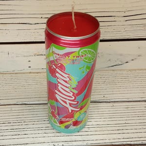 May include: A red candle in a can-shaped container with a cherry twist design. The can features the text "Alani" in a colorful, stylized font, along with cherry and lime graphics. The candle has a white wick.